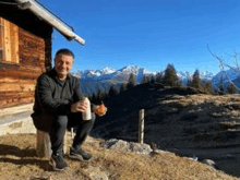 a man is sitting on a stump in front of a wooden cabin holding a bottle of beer .