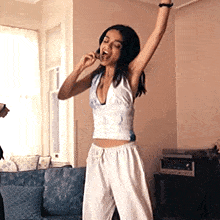 a woman in a white crop top and white pants is standing in a living room with her arms in the air