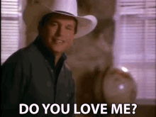 a man wearing a cowboy hat is standing in front of a window and saying `` do you love me '' .