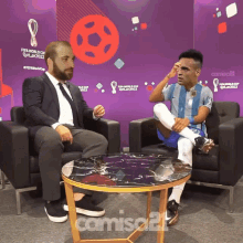 a man in a suit sits next to a man in a soccer uniform in front of a fifa world cup qatar 2022 sign