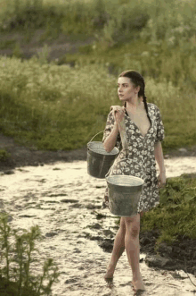 a woman in a dress is walking through a stream carrying two buckets of water .