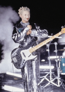 a woman in a silver outfit is holding a guitar and singing into a microphone