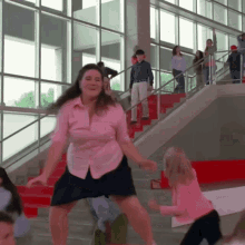 a woman in a pink shirt and black skirt is dancing on the stairs in front of a crowd .