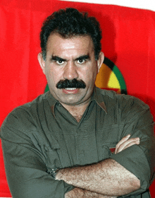 a man with a mustache stands with his arms crossed in front of a red flag