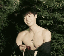 a shirtless man wearing a cross necklace is smiling in front of trees