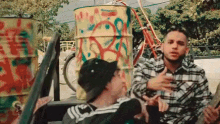 a man in a plaid shirt is sitting in the back of a truck next to a barrel with graffiti on it .