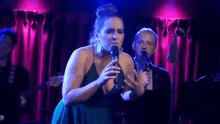 a woman is singing into a microphone on a stage while a man stands behind her .