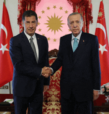 two men in suits shaking hands in front of a flag