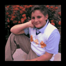 a young boy wearing a blue shirt and a white vest is sitting in front of a bush of red roses