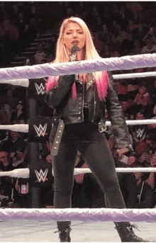 a woman in a leather jacket stands in a wrestling ring with a microphone in her hand