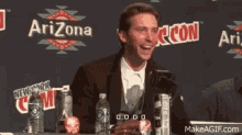 a man sitting at a table with bottles of water in front of a sign that says arizona new york comic con