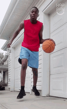 a young man in a red tank top and blue shorts is holding a basketball in front of a garage door