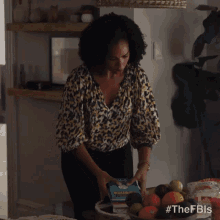 a woman in a leopard print shirt is standing at a table with a bowl of fruit and a bottle of milk .