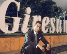 a man in a suit sits on a bench in front of a large sign that says giresun