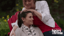 two women are sitting on a red chair and one is putting her hand on the other 's forehead ..