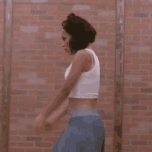 a woman in a white crop top and blue pants is standing in front of a brick wall .