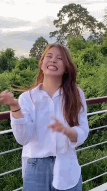 a woman in a white shirt and blue jeans is standing on a balcony with trees in the background .