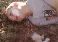 a boy with a bandage on his face is laying in the grass wearing a t-shirt that says rvtyi