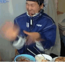 a man in a blue jacket is sitting at a table with plates of food and a bowl of food .