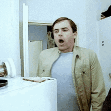 a man in a tan jacket is yawning in front of a white refrigerator .