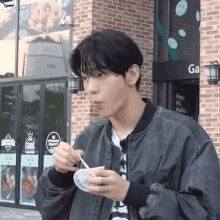 a young man in a bomber jacket is eating ice cream with a spoon in front of a brick building .