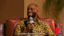 a man sitting on a red couch holding a microphone with the words " and in ghana we say "