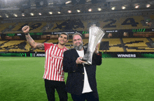 a man in a red and white striped shirt holds a trophy next to another man