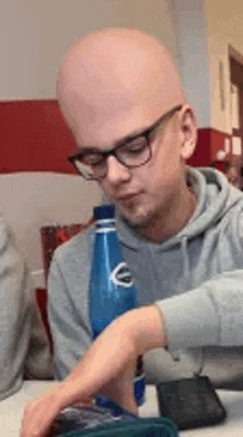 a bald man with glasses is sitting at a table with a bottle of water and a cell phone .