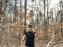 a man wearing sunglasses and a hat stands in the woods holding a stick