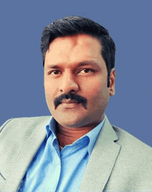 a man with a mustache wearing a suit and a blue shirt is sitting in front of a blue background .