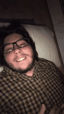 a man with glasses and a plaid shirt smiles while laying on a bed