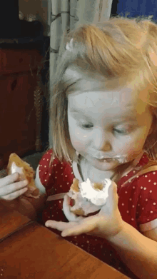 a little girl in a red and white polka dot dress is eating a donut with whipped cream on her face