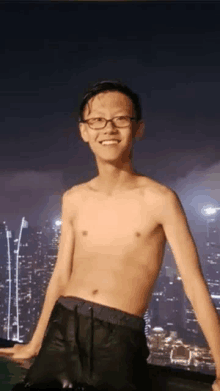 a young man without a shirt and glasses is standing in front of a city skyline at night .