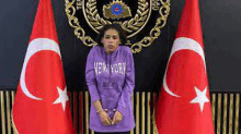 a woman in a purple new york sweatshirt is standing in front of two flags .