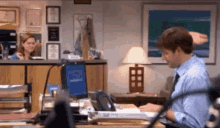 a man in a blue shirt and tie is sitting at a desk in front of a computer monitor .