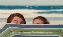 two girls peeking out from behind a beach umbrella with the words look at him sitting up there being gorgeous