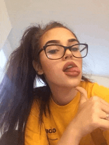 the girl is wearing glasses and a yellow shirt and sticking her tongue out .