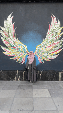 a woman in a hijab stands in front of a painting of wings