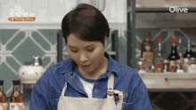 a woman wearing an apron and a denim shirt is standing in front of a shelf of olive bottles .