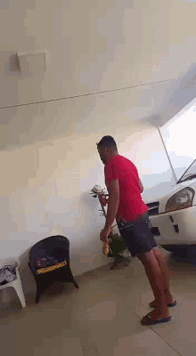 a man in a red shirt and black shorts is standing in front of a white car in a garage .