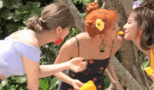 three women are standing next to each other in a garden and one of them is eating an orange .