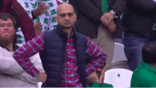 a man in a plaid shirt and vest is sitting in a crowd with his hands on his hips .