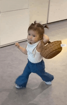 a little girl in a white tank top and blue pants is walking with a basket on her back .