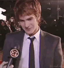 a man in a suit and tie is talking into a microphone on a red carpet .