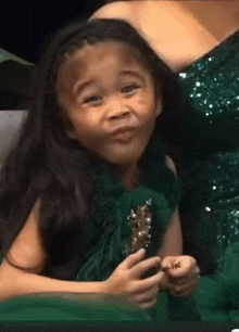 a little girl in a green dress is making a funny face while holding a trophy .