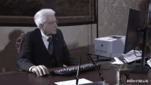 a man in a suit sits at a desk with a computer and a clock that has ask news on it