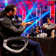 a man sitting in a chair talking into a microphone while a woman holds a wwe trophy in front of him