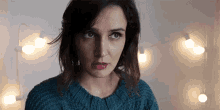 a woman in a blue sweater looks at the camera with a string of lights behind her