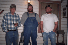 three men standing in front of a window one of whom is wearing overalls and a shirt that says john deere on it