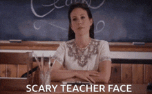 a woman is sitting at a desk in front of a chalkboard with the words scary teacher face written on it .